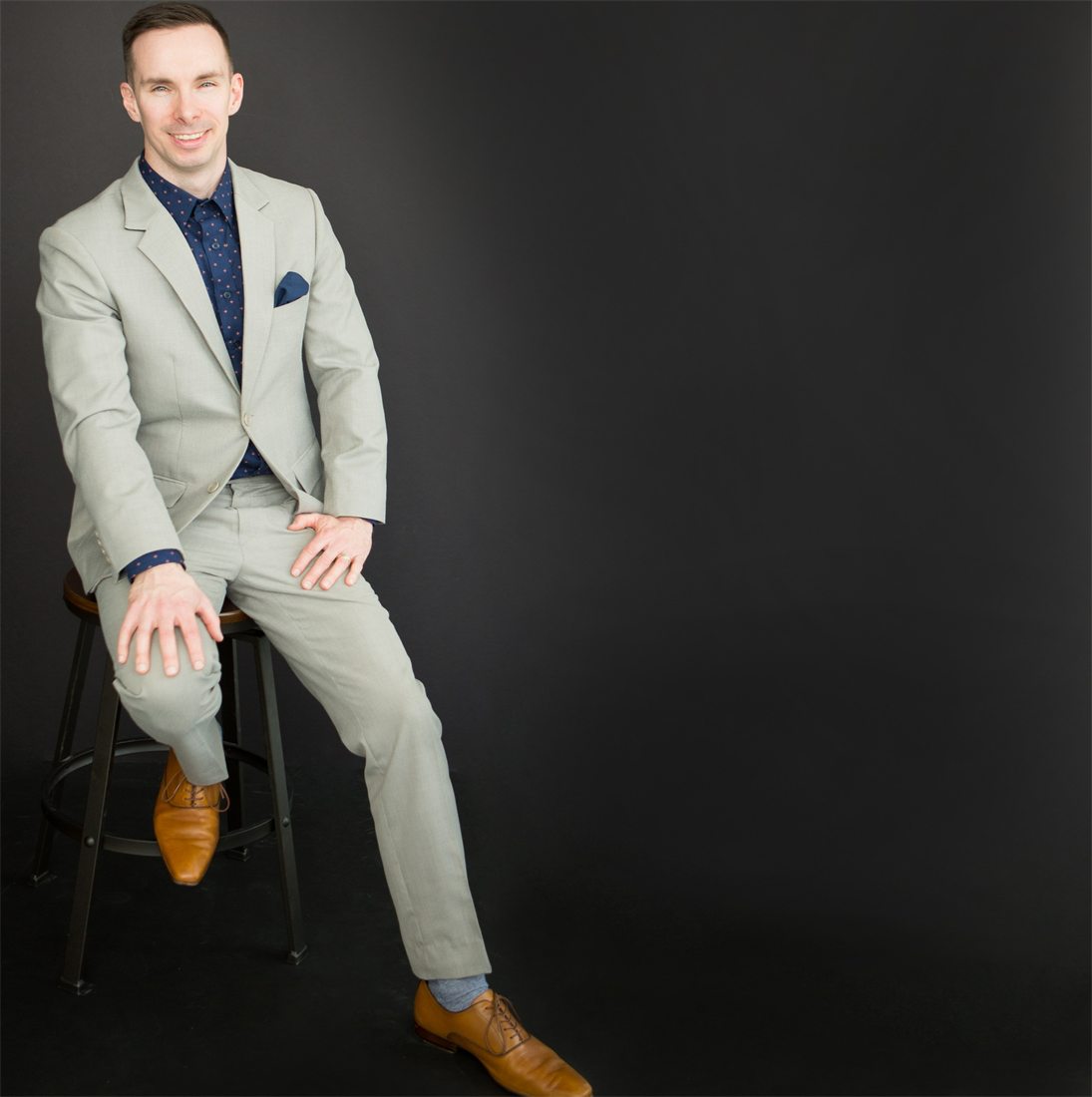 Derek seated in a light grey made-to-measure suit with a navy patterned shirt, navy pocket square and tan leather shoes, relaxed and at ease