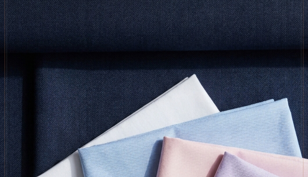 Five folded dress shirts in white, light blue, pink, lavender and grey, fanned across a navy herringbone suit fabric