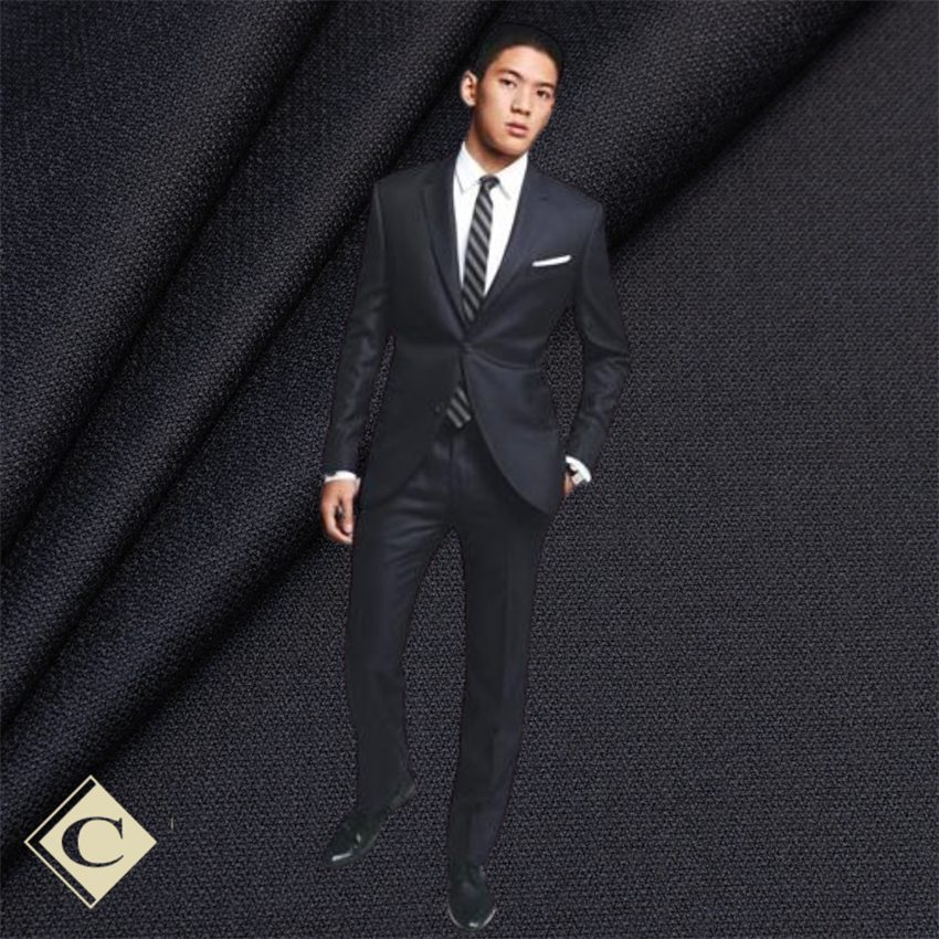 A man in a charcoal grey custom suit with a white shirt and striped tie, against a charcoal fabric backdrop