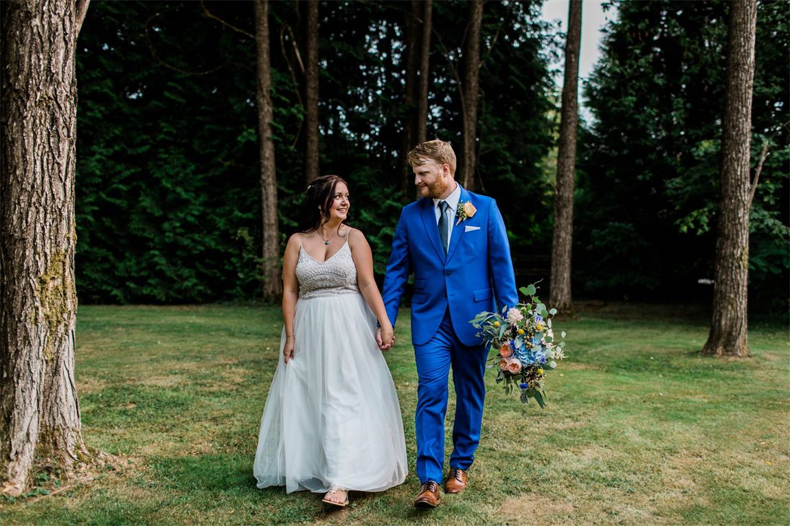 A groom in a bright blue made-to-measure suit with a boutonniere and bouquet, walking hand in hand with his bride on the lawn