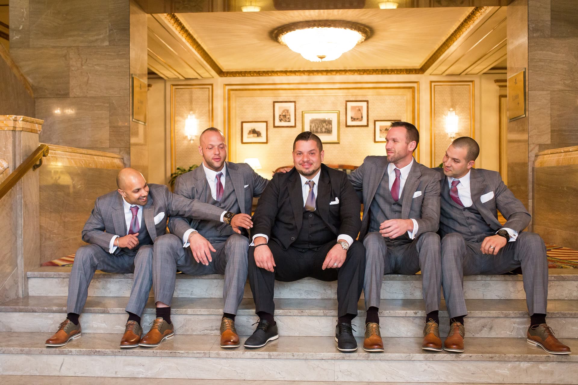 A groom in a charcoal three-piece suit seated with four groomsmen in grey suits and burgundy ties in an elegant lobby