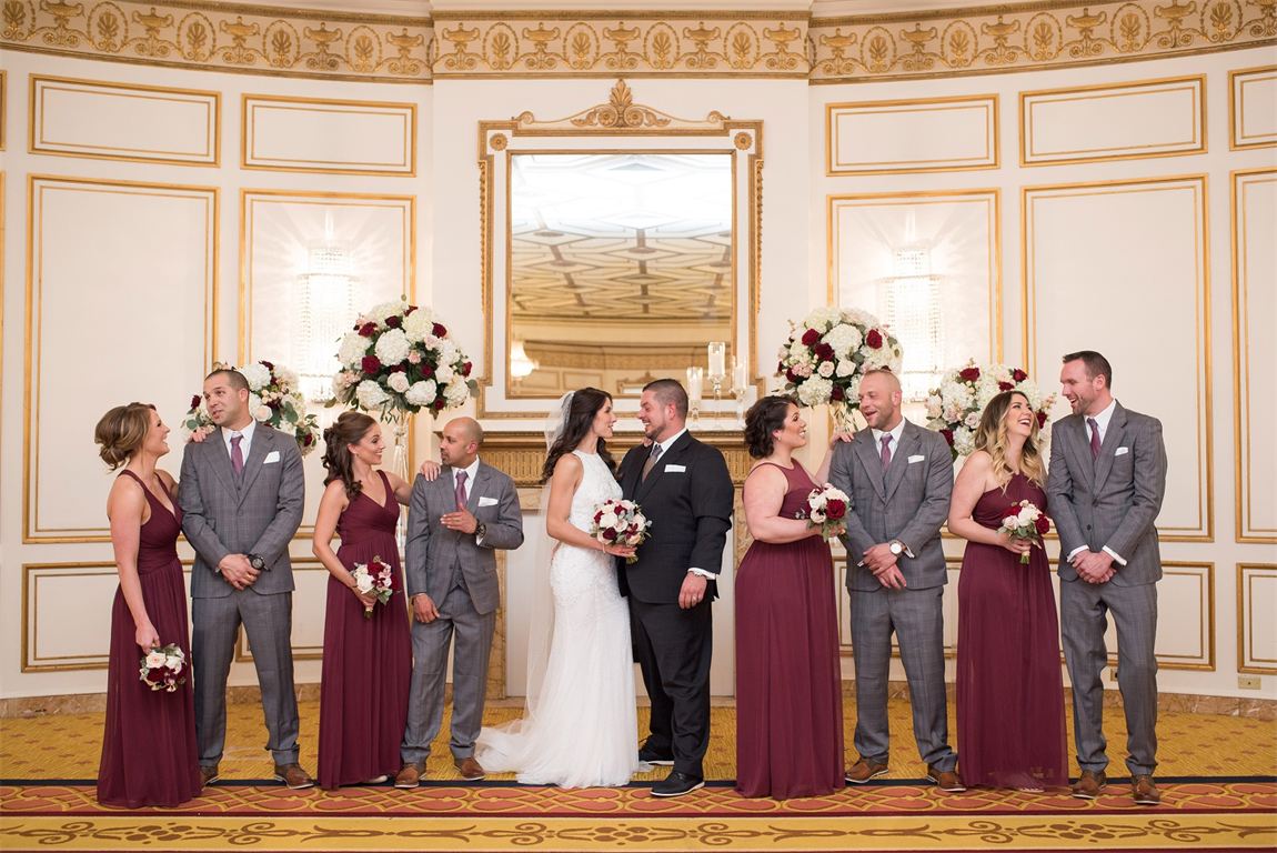 A full wedding party in a gilded ballroom: the groom and bride centred, groomsmen in grey suits and bridesmaids in burgundy, each suit built to fit and coordinated across the group
